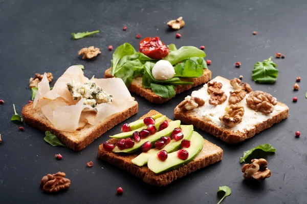 Broodjes met een verscheidenheid van toppings zoals rucola, gedroogde tomaten, zachte kaas, blauwe kaas, noten en avocado's op een donkere achtergrond. Het concept van gezonde vegetarische voeding. — Stockfoto