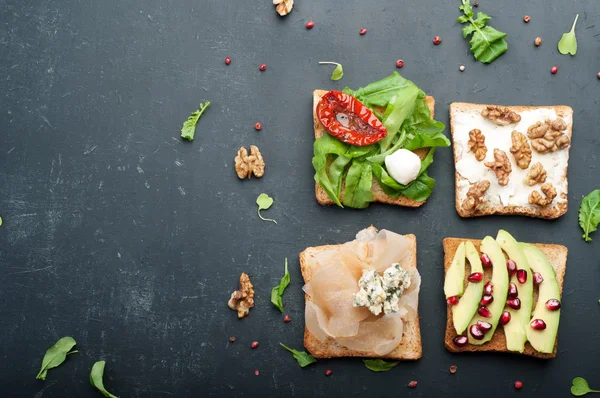 Sandwiches mit verschiedenen Belägen wie Rucola, getrockneten Tomaten, Weichkäse, Blauschimmelkäse, Nüssen und Avocados auf dunklem Hintergrund. das Konzept der gesunden vegetarischen Ernährung. — Stockfoto