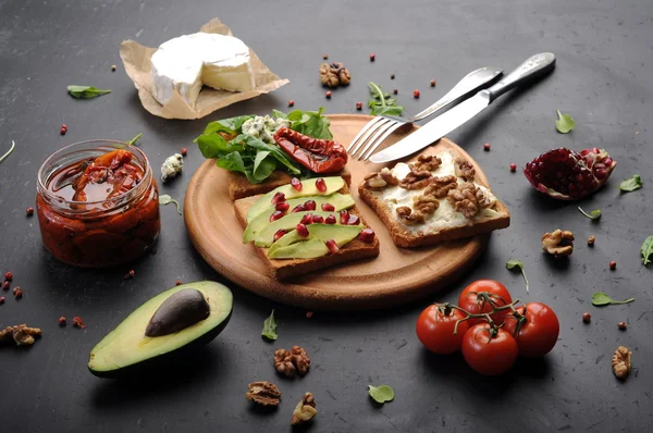 Sandwiches mit verschiedenen Belägen wie Rucola, getrockneten Tomaten, Weichkäse, Blauschimmelkäse, Nüssen und Avocados auf dunklem Hintergrund. das Konzept der gesunden vegetarischen Ernährung. — Stockfoto