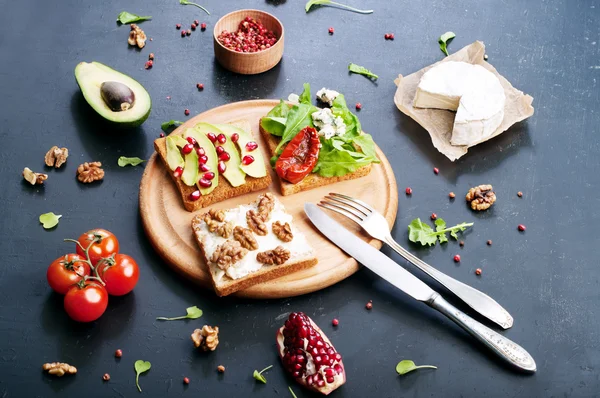 Sandwiches with a variety of toppings such as arugula, dried tomatoes, soft cheese, blue cheese, nuts and avocados on a dark background. The concept of healthy vegetarian food. — Stock Photo, Image