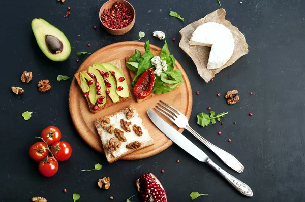 Sandwiches with a variety of toppings such as arugula, dried tomatoes, soft cheese, blue cheese, nuts and avocados on a dark background. The concept of healthy vegetarian food. — Stock Photo, Image