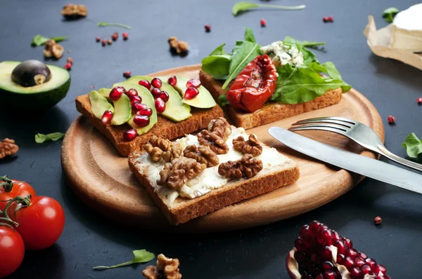 Sandwiches mit verschiedenen Belägen wie Rucola, getrockneten Tomaten, Weichkäse, Blauschimmelkäse, Nüssen und Avocados auf dunklem Hintergrund. das Konzept der gesunden vegetarischen Ernährung. — Stockfoto