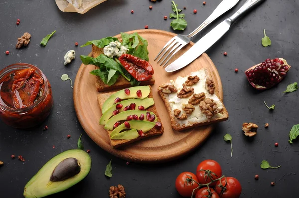 Sandwiches mit verschiedenen Belägen wie Rucola, getrockneten Tomaten, Weichkäse, Blauschimmelkäse, Nüssen und Avocados auf dunklem Hintergrund. das Konzept der gesunden vegetarischen Ernährung. — Stockfoto