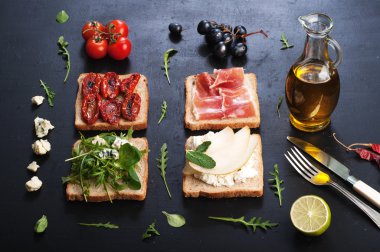 Sandwiches with a variety of toppings on a dark surface. Sandwiches with arugula, leaf mash, blue cheese, sun-dried tomatoes, slices of pear and ham. The concept of useful home cooking
