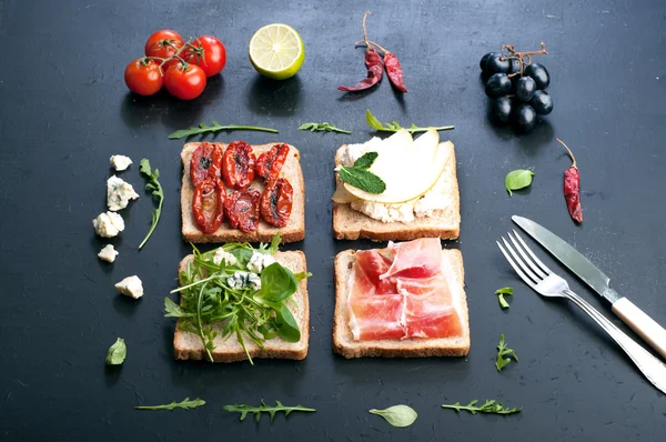 Sandwiches with a variety of toppings on a dark surface. Sandwiches with arugula, leaf mash, blue cheese, sun-dried tomatoes, slices of pear and ham. The concept of useful home cooking
