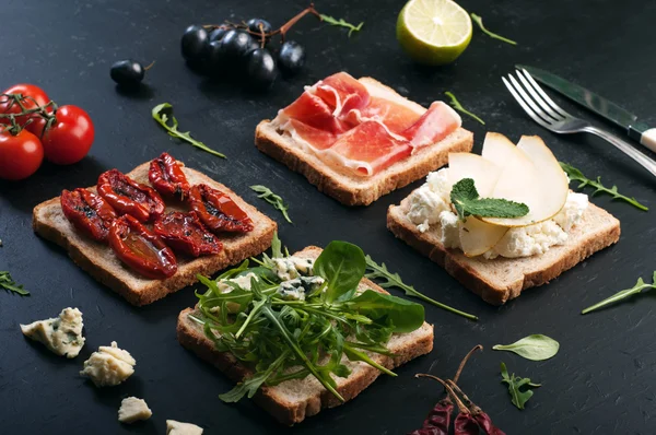 Sandwiches mit verschiedenen Belägen auf dunkler Oberfläche. Sandwiches mit Rucola, Blattpüree, Blauschimmelkäse, sonnengetrockneten Tomaten, Birnen- und Schinken-Scheiben. das Konzept der nützlichen Hausmannskost — Stockfoto