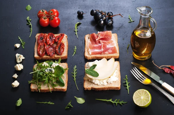 Sandwiches with a variety of toppings on a dark surface. Sandwiches with arugula, leaf mash, blue cheese, sun-dried tomatoes, slices of pear and ham. The concept of useful home cooking — 스톡 사진