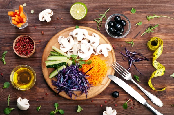The concept of dietetic vegetarian food. Bright juicy shredded vegetables, such as carrots, purple cabbage, mushrooms and cucumbers, which lies on a circular wooden cutting board. Natural organic products, ready to eat — Stock Photo, Image