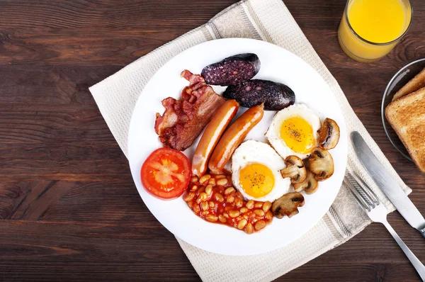 O café da manhã clássico inglês com ovos fritos, salsichas, pudim preto, cogumelos fritos e feijão com tomates em um fundo de madeira marrom. O conceito de um café da manhã saudável . — Fotografia de Stock