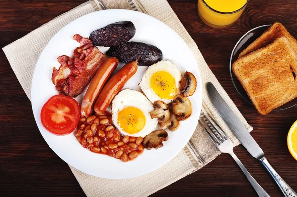 经典的英式早餐，包括煎蛋、香肠、黑布丁、炸蘑菇和带番茄的棕色木制背景。有益早餐的概念. — 图库照片