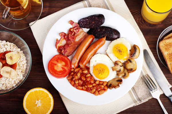 Классический английский завтрак с жареными яйцами, сардельками, черным пудингом, жареными грибами и фасолью с помидорами на коричневом деревянном фоне. Концепция здорового завтрака
.