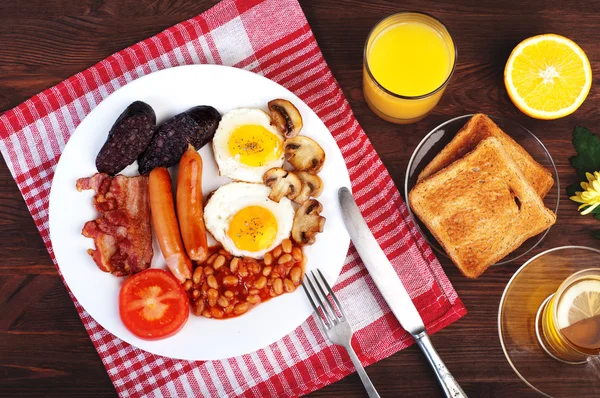 La classica colazione all'inglese con uova fritte, salsicce, budino nero, funghi fritti e fagioli con pomodori su uno sfondo di legno marrone. Il concetto di una colazione sana . — Foto Stock