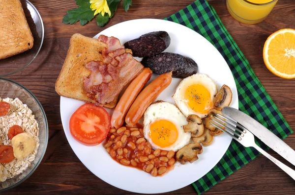 O café da manhã clássico inglês com ovos fritos, salsichas, pudim preto, cogumelos fritos e feijão com tomates em um fundo de madeira marrom. O conceito de um café da manhã saudável . — Fotografia de Stock
