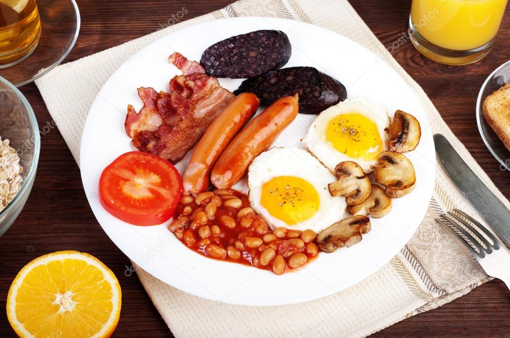 The classic English breakfast with fried eggs, sausages, black pudding, fried mushrooms and beans with tomatoes on a brown wooden background. The concept of a wholesome breakfast.