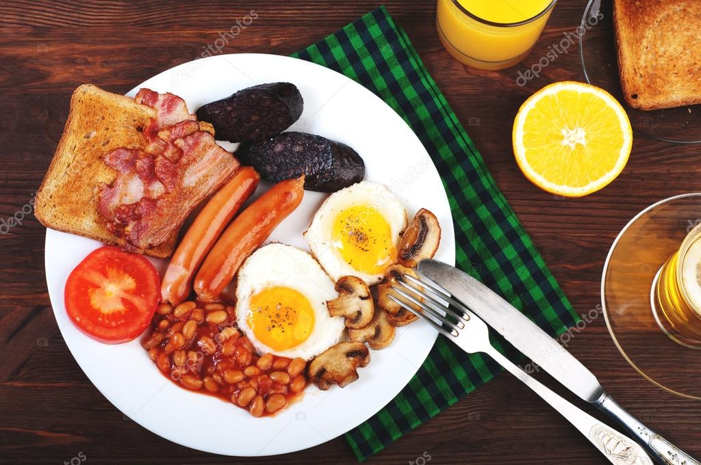 The classic English breakfast with fried eggs, sausages, black pudding, fried mushrooms and beans with tomatoes on a brown wooden background. The concept of a wholesome breakfast.