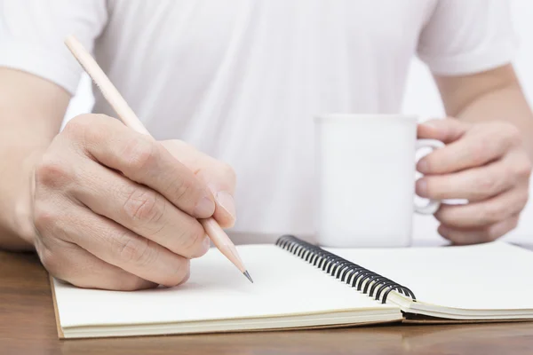 Hombre escribiendo en el cuaderno y sosteniendo una taza de bebida caliente — Foto de Stock