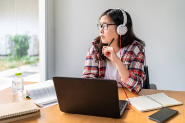 Kulaklık takan genç Asyalı kız öğrenci Corona virüsünün yayılması nedeniyle şehir tecridi sırasında çalışırken ve çalışırken pencereden dışarı bakıyor.