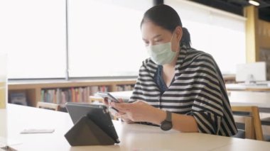 Tıbbi maske takan genç Asyalı kadın internet, teknoloji veya internet iş konseptine bağlanmak için telefon kullanıyor.