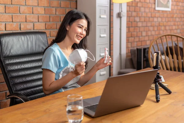 Happy young Asian businesswoman holding N95 face mask and alcohol spray bottle to show her social media fan page about stay safe during COVID lockdown through her online channel on her phone and computer from her living room of her house