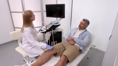 Beyaz önlüklü tıp uzmanı erkek hastanın dizinde ultrason uygular ve ultrasonik muayenede yaralanmayı değerlendirir. Diz ultrasonu