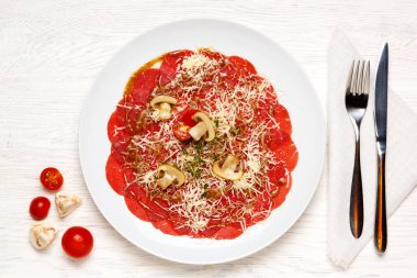Biftek carpaccio güzel bir şekilde beyaz bir tabakta servis edilir ve domates, şampiyon ve peynirle süslenir. Üst görünüm