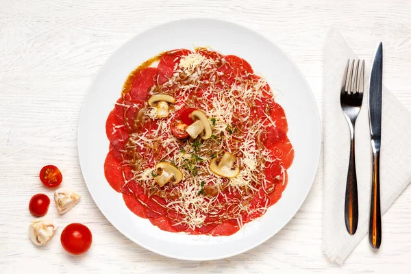 Biftek carpaccio güzel bir şekilde beyaz bir tabakta servis edilir ve domates, şampiyon ve peynirle süslenir. Üst görünüm