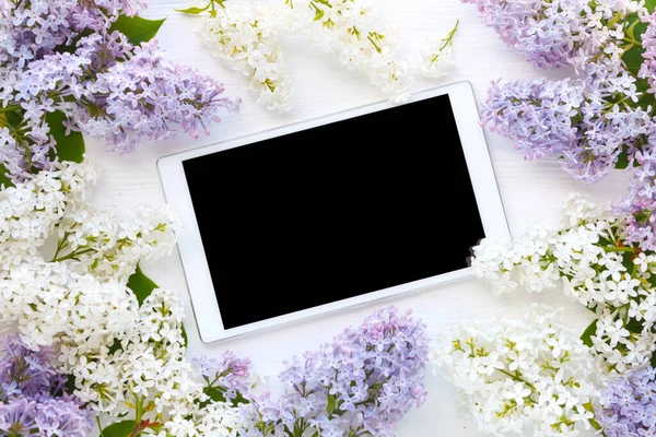 White tablet computer and lilac flowers on white wooden background. Top view. Flat lay. Copy space