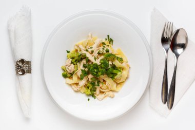 Brokolili makarna ve çatal ve kaşıkla beyaz ahşap bir masada beyaz bir tabakta tavuk. Üst görünüm