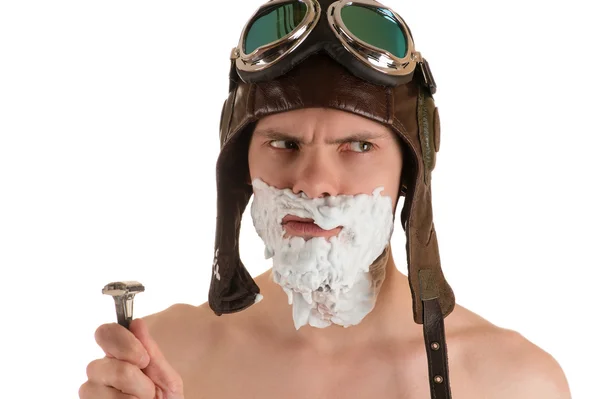 man with shaving foam on his face in flight helmet and flying goggles and razor in his hand looking away