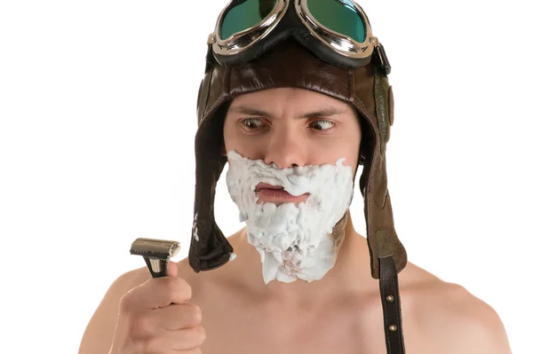 man with shaving foam on his face in flight helmet and flying goggles looking at razor in his hand