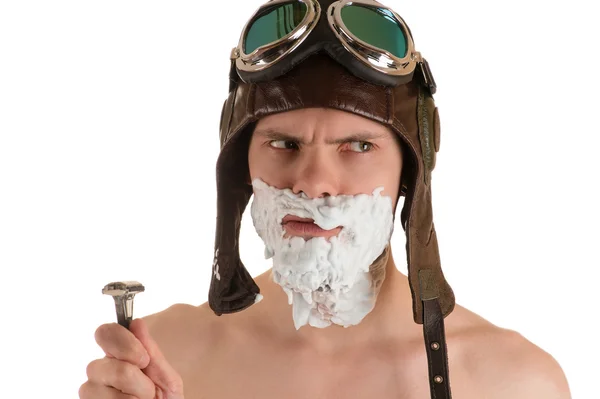 Hombre con espuma de afeitar en su cara en casco de vuelo y gafas voladoras y maquinilla de afeitar en su mano mirando hacia otro lado Imagen de archivo