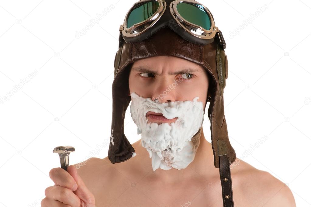man with shaving foam on his face in flight helmet and flying goggles and razor in his hand looking away