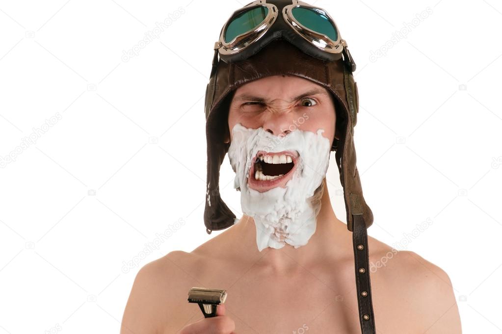 screaming man with narrowed eyes with shaving foam on his face in flight helmet and flying goggles with razor in his hand