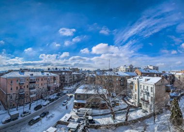 Kış kenti sokakları bulutlarla dolu. Karla kaplı çatılar ve arabalarla dolu bir yol. Mavi, soğuk bir gökyüzüne karşı bir bulut manzarası..