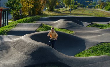Şehir parkında eğri büğrü asfalt tulumba pistinde scooter süren odaklanmış bir çocuk. Aktif çocukluk, denge eğitimi, açık hava eğlencesi ve sportif yaşam tarzı