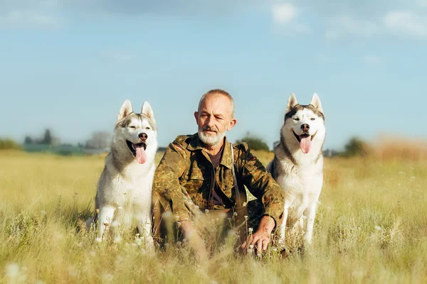 Viejo con barba sentado en un pajar con sus perros, disfrutando del atardecer de verano. Husky siberiano —  Fotos de Stock