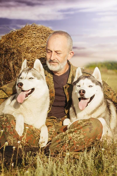 Viejo con barba sentado en un pajar con sus perros, disfrutando del atardecer de verano . —  Fotos de Stock