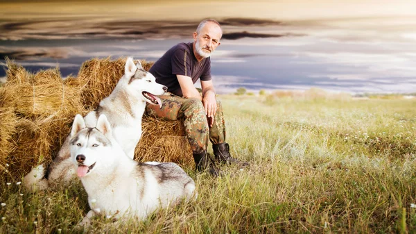 Viejo con barba sentado en un pajar con sus perros, disfrutando del atardecer de verano . —  Fotos de Stock