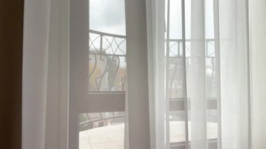 Açık beyaz pencere örtüsünün dokusu, şehir mimarisi ve mevsimsel doğanın bulanık arka planıyla, yumuşak sabah ışığının oturma odasına girmesine olanak sağlıyor.. 