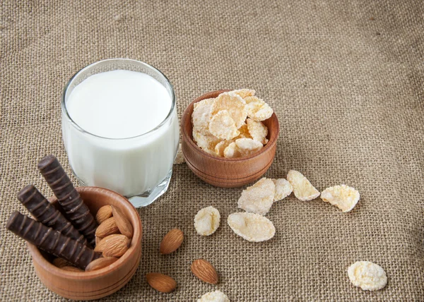 Un vaso de leche con nueces de almendras, hojuelas de maíz, chocolates, sobre un fondo de saqueo, arpillera , —  Fotos de Stock