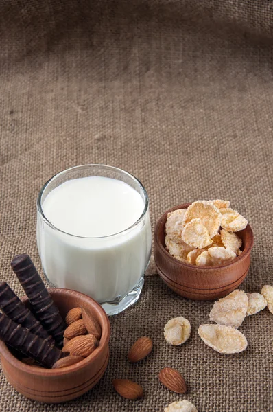 Un vaso de leche con almendras, hojuelas de maíz, chocolates, sobre fondo de arpillera — Foto de Stock