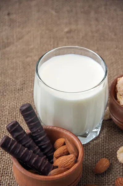 Un vaso de leche con almendras, hojuelas de maíz, chocolates, sobre fondo de arpillera —  Fotos de Stock