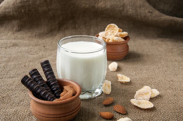 Un vaso de leche con almendras, hojuelas de maíz, chocolates, sobre fondo de arpillera —  Fotos de Stock