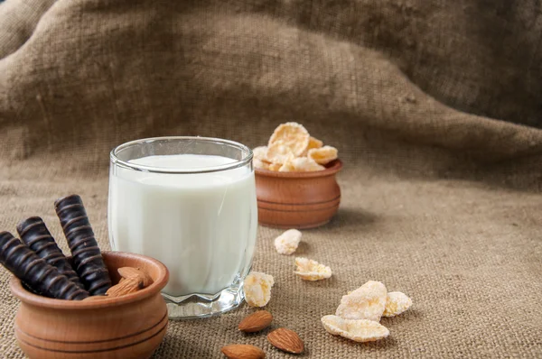 Un vaso de leche con almendras, hojuelas de maíz, chocolates, sobre fondo de arpillera —  Fotos de Stock