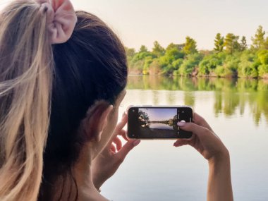 Athalassa Parkı, Kıbrıs - 9 Ekim 2020: Bir kız güzel güneşli bir sonbahar öğleden sonrasında Athalassa doğal parkında cep telefonuyla gölün fotoğrafını çekiyor.