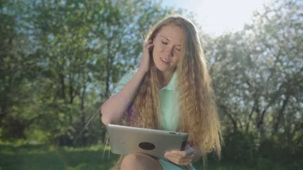 Chica con pelo rojo largo mira a la tableta en el jardín de manzanas — Vídeo de stock