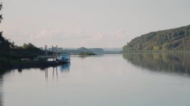 Çerçevede, kamera nehri gören bir manzarayla soldan sağa hareket ediyor. Solda teknesi olan bir iskele var. Nehrin karşı kıyısında yeşil bitki örtüsü olan dik bir banka var. Gün batımı zamanı.