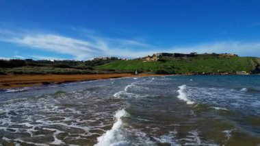 Gozo 'daki en güzel Malta sahilinin hava aracı görüntüsü, berrak mavi suyu ve yemyeşil bitki örtüsüyle büyüleyici dalgaları olan canlı kırmızı Ramla plajı ve güneşli bir günde kont sabahında ünlü Mixta Mağarası arka planı.