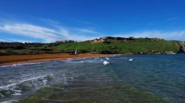 Gozo 'daki en güzel Malta sahilinin hava aracı görüntüsü, berrak mavi suyu ve yemyeşil bitki örtüsüyle büyüleyici dalgaları olan canlı kırmızı Ramla plajı ve güneşli bir günde kont sabahında ünlü Mixta Mağarası arka planı.