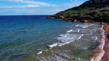 Gozo 'daki en güzel Malta sahilinin hava aracı görüntüsü, berrak mavi suyu ve yemyeşil bitki örtüsüyle büyüleyici dalgaları olan canlı kırmızı Ramla plajı ve güneşli bir günde kont sabahında ünlü Mixta Mağarası arka planı.